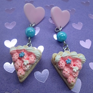 May include: A pair of pink heart dangle earrings with a blue bead and a pink and white strawberry pie charm.