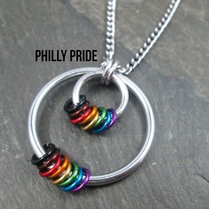 May include: A silver necklace with a circle pendant featuring a cluster of rainbow-colored rings. The pendant is inscribed with the text "PHILLY PRIDE".