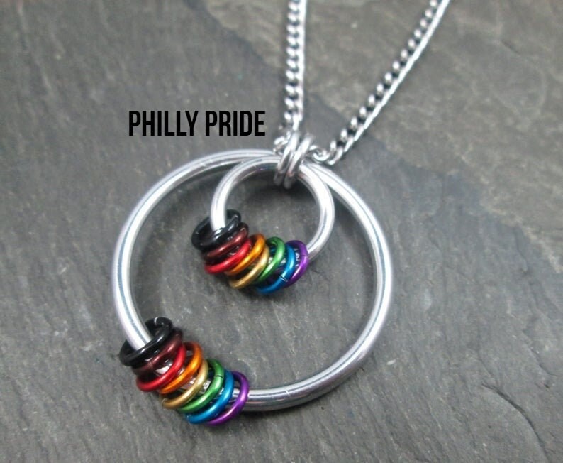 May include: A silver necklace with a circle pendant featuring a cluster of rainbow-colored rings. The pendant is inscribed with the text "PHILLY PRIDE".