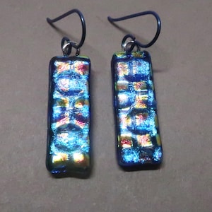 May include: A pair of rectangular dichroic glass earrings. The earrings have a blue and iridescent color scheme with hints of red and yellow. The ear wires are dark blue. The earrings are approximately 1 inch long.
