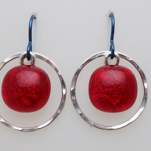May include: A pair of earrings featuring a red, glittery, round bead suspended within a hammered silver-tone circle. The earrings have blue ear wires, creating a striking contrast. The jewelry is displayed against a plain white background.