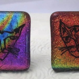 May include: Two square glass cabochons featuring black line art of cat faces. The left piece displays a vibrant iridescent finish with streaks of blue, purple, and green. The right piece features a warm iridescent finish with orange, red, and gold tones.