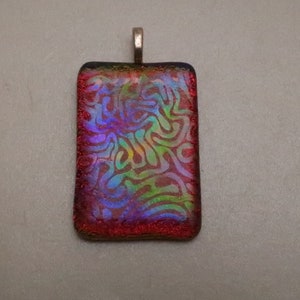 May include: Rectangular pendant with a silver-colored bail. The pendant features a vibrant, iridescent design with swirling patterns in shades of red, purple, green, and yellow. The edges are a deep red color.