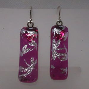 May include: A pair of magenta-colored rectangular earrings with silver leaf-like designs. The earrings have a silver hook closure. The earrings are a unique accessory.