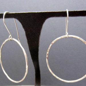 May include: A pair of silver hoop earrings with a hammered texture. The earrings are hanging from a black display stand.