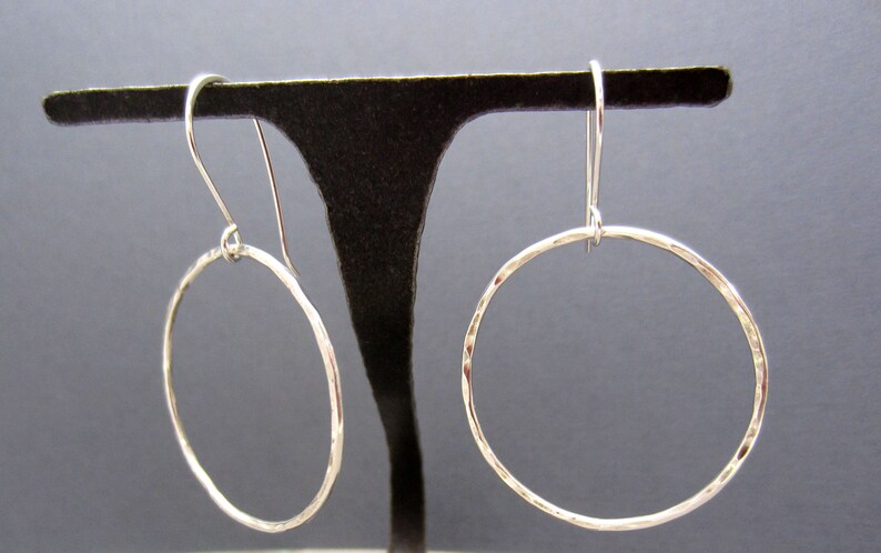 May include: A pair of silver hoop earrings with a hammered texture. The earrings are hanging from a black display stand.