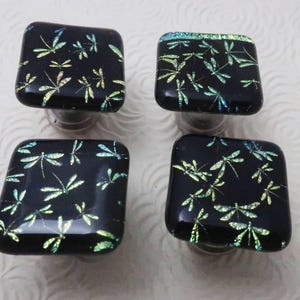 May include: Four square cabinet knobs made of black glass, each featuring a repeating pattern of iridescent green and blue dragonfly silhouettes. The knobs have rounded edges and a smooth, glossy finish, with a metallic base visible underneath.