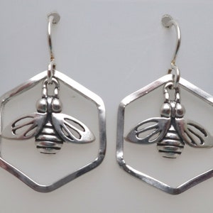 May include: A pair of silver-toned bee earrings. Each earring features a detailed bee charm suspended within a hexagonal frame. The earrings have a hook closure for pierced ears. The design is simple and elegant.