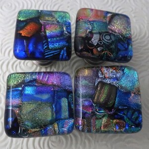 May include: Four square cabinet knobs made of dichroic glass. Each knob features a mosaic-like pattern with iridescent shades of blue, green, purple, and orange. The glass surfaces are smooth and reflective, displaying a multi-colored metallic sheen.