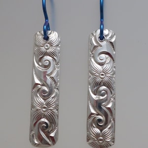 May include: A pair of silver rectangular earrings with a floral and swirl design. The earrings have blue ear wires. The floral pattern is carved into the silver, creating a textured surface. The earrings are displayed against a light background.