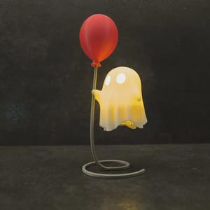 Könnte beinhalten: Eine kleine, beleuchtete geisterförmige Lampe mit sanftem weißem Licht und zwei kreisförmigen Augen. Der Geist ist an einem dünnen, geschwungenen Metallständer befestigt, der einen roten Ballon hält. Der Fuß des Ständers ist eine flache Metallspirale.