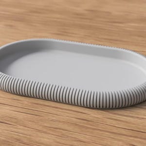 Könnte beinhalten: Ein hellgraues, ovales Tablett mit geripptem Rand. Das Tablett besteht aus einem festen Material und liegt auf einer Holzoberfläche. Das Design ist schlicht und modern, für verschiedene Zwecke geeignet.
