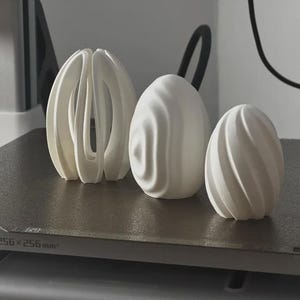 Könnte beinhalten: Drei weiße 3D-gedruckte Objekte mit unterschiedlichen geometrischen Designs. Das linke Objekt hat eine offene, käfigartige Struktur. Das mittlere Objekt hat eine gewellte, organische Oberfläche. Das rechte Objekt zeigt ein Spiralmuster. 256x256x256mm.