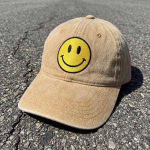 Op de afbeelding: Een beige honkbalpet met een verweerde, verwassen stofafwerking. Het voorpaneel is voorzien van een ronde geborduurde patch met een felgeel smileygezicht, bestaande uit twee zwarte ovale ogen en een brede, gebogen zwarte mond.