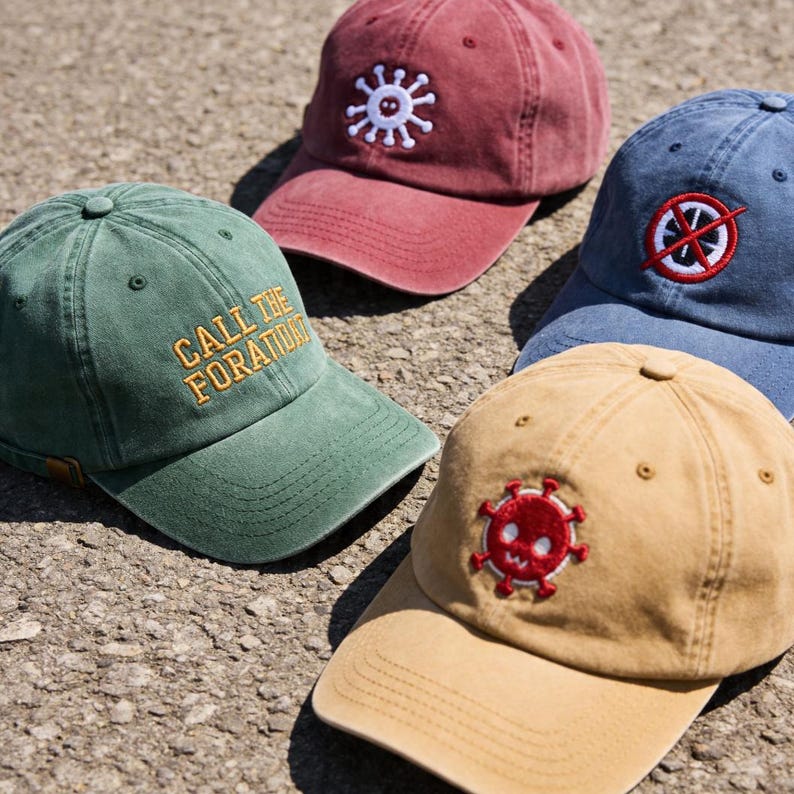 Op de afbeelding: Vier baseballpetten in groen, rood, blauw en beige. De groene pet heeft de gele geborduurde tekst CALL THE FORATIDAT. De rode pet heeft een wit virusicoon, de blauwe pet toont een rode cirkel met een streep erdoor en de beige pet een rood virusicoon.