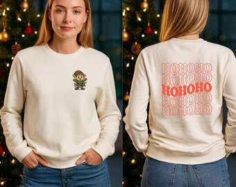 May include: Cream-coloured sweatshirt with an embroidered elf on the front. The back of the sweatshirt has the text "HOHOHO" in a repeating pattern in red and pink. The model is wearing blue jeans.