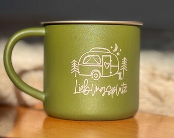 Op de afbeelding: Een groene campingmok met een wit ontwerp van een camper, bomen, een halve maan en sterren. De tekst "Lieblingsplatz" staat onder de camper.