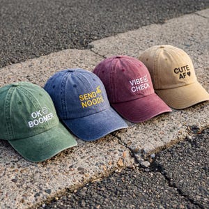 Op de afbeelding: Vier baseballpetten in groen, blauw, bordeaux en beige. De groene pet heeft de tekst OK BOOMER met een smiley. De blauwe pet heeft de tekst SEND NOODS met een noedelkom-icoon. De bordeaux pet heeft de tekst VIBE CHECK met een vinkje. De beige pet heeft de tekst CUTE AF met een hartje.