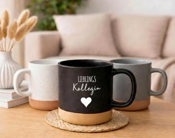 Op de afbeelding: Drie keramische mokken in wit, grijs en zwart, elk met een bruine basis. De zwarte mok heeft de tekst "LIEBLINGS Kollegin" en een wit hart. De mokken staan op een houten tafel.