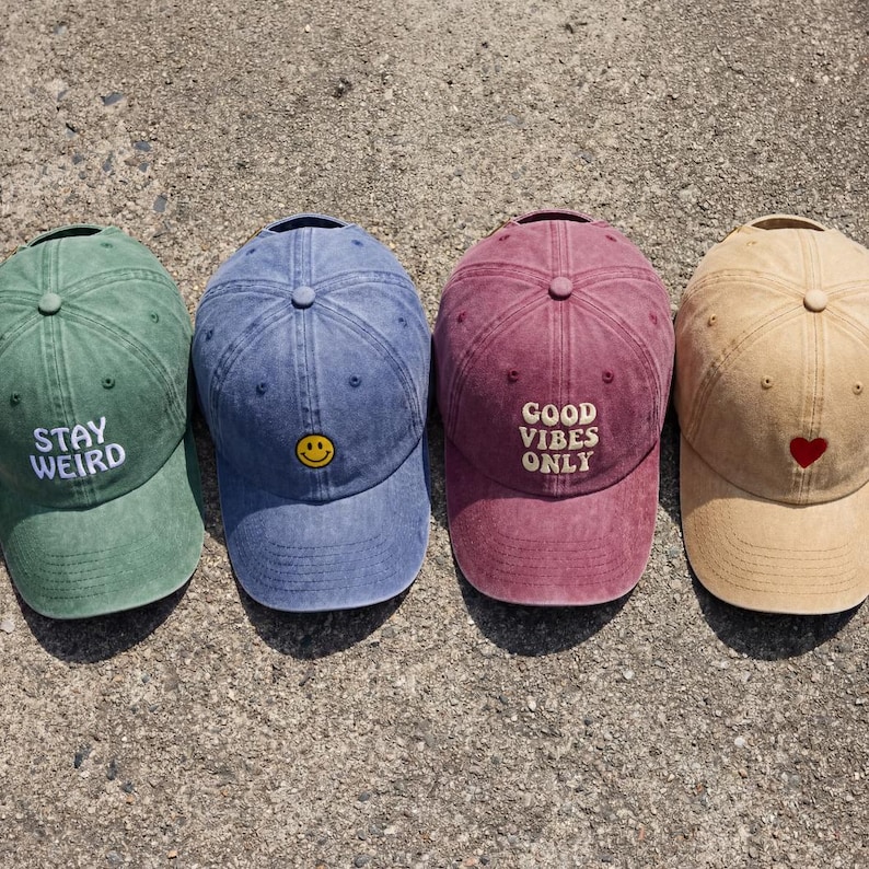 Op de afbeelding: Vier baseballpetten in groen, blauw, bordeauxrood en beige. De groene pet heeft de witte tekst STAY WEIRD. De blauwe pet heeft een geel smileygezicht. De bordeauxrode pet toont de witte tekst GOOD VIBES ONLY. De beige pet heeft een rood hart.