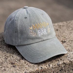Könnte beinhalten: Graue Baseballkappe mit dem gestickten Text "MAMA BÄR" in Gold und Weiß. Die Kappe hat einen gebogenen Schirm und einen Stoffriemenverschluss. Das Design umfasst Herz- und Blätterakzente.
