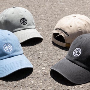 Op de afbeelding: Een collectie van vier baseballpetten in lichtgrijs, lichtblauw, beige en antraciet. De grijze pet heeft een rond logo, de blauwe pet een rond embleem en de beige pet toont de tekst VOLGA OH MOOIST.