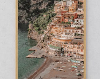 May include: A framed print featuring a coastal Italian town. The image showcases colorful buildings cascading down a cliffside to a beach with boats. The water is turquoise, and the sky is overcast. The frame is a light wood color.