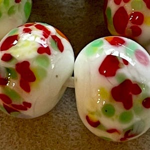 May include: Two oval-shaped glass beads featuring a white base with a speckled pattern of red, light green, and yellow spots. The beads have a smooth, glossy finish and are arranged side by side, showing a reflective surface and rounded edges.