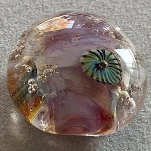 May include: A round, clear glass paperweight featuring internal swirls of soft pink and amber hues. The surface is embedded with numerous small air bubbles and a single, prominent, spiraled teal and black glass accent piece.