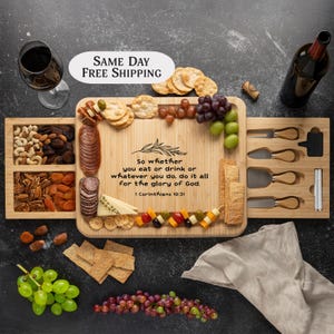May include: A wooden serving board with a sliding drawer for cheese knives. The board has a quote from the Bible, "So whether you eat or drink or whatever you do, do it all for the glory of God. 1 Corinthians 10:31". The board is filled with various snacks, including grapes, crackers, cheese, and nuts.