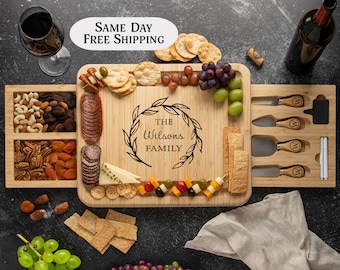 May include: A wooden serving board with a sliding drawer, featuring a personalized inscription "THE Wilsons FAMILY" in a wreath design. The board is filled with various snacks, including cheese, crackers, grapes, and nuts. The board also includes a set of cheese knives and a small chalkboard.