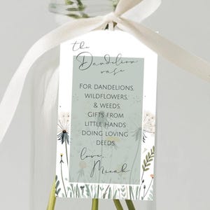 Puede incluir: Un jarrón de cristal transparente contiene dientes de león amarillos y blancos. Una cinta blanca está atada alrededor del cuello del jarrón. Una etiqueta con texto está adjunta al jarrón. La etiqueta dice "el jarrón de diente de león" y otro texto.