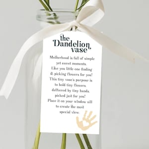 Puede incluir: Un jarrón de cristal transparente con una cinta blanca y una etiqueta que dice "the Dandelion vase". El jarrón contiene dientes de león amarillos y otras flores pequeñas. La etiqueta tiene texto sobre la maternidad y las flores.