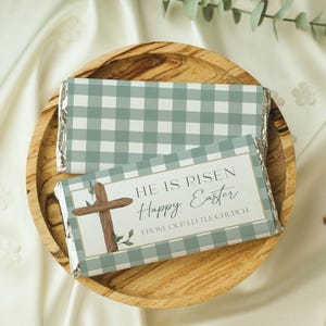 Puede incluir: Dos barras de chocolate con envoltorios de papel de aluminio plateado, con un patrón de cuadros vichy verde y blanco y texto temático de Pascua. Una barra muestra una cruz de madera y las palabras "He Is Risen Happy Easter."