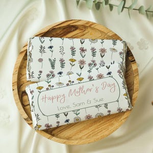 May include: Two chocolate bars wrapped in white paper with a floral pattern. The top bar has a pink floral pattern. The bottom bar has a pink floral pattern and the text "Happy Mother's Day Love. Sam & Sue".