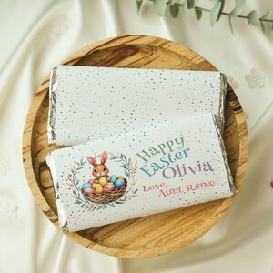 May include: Two white chocolate bars wrapped in foil with a silver glitter design. The top bar has a bunny rabbit holding colourful Easter eggs in a basket. The bottom bar has the text "Happy Easter Olivia Love, Aunt Renee".