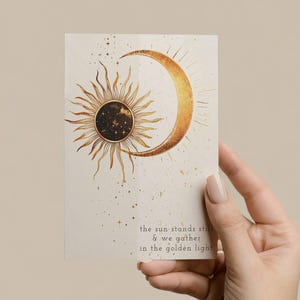 May include: A rectangular card featuring a celestial design with a sun and crescent moon in gold tones. The sun has radiating lines, and the moon is curved. The text reads: "the sun stands still & we gather in the golden light."