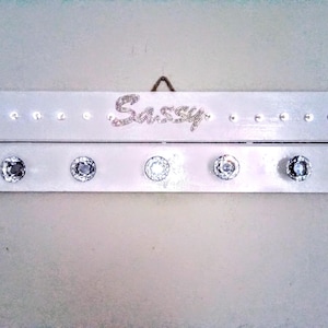 May include: A white wooden wall-mounted organizer with the word "Sassy" spelled out in rhinestones. It features a row of small, clear rhinestones along the top and five large, clear, decorative knobs below.