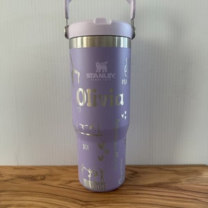 Peut inclure: Une bouteille d'eau en acier inoxydable de couleur violette avec une poignée de transport. La bouteille a un motif argenté avec les mots "Stanley", "Olivia", "Peace", "Gymnastics" et "10!" La bouteille est posée sur une surface en bois.