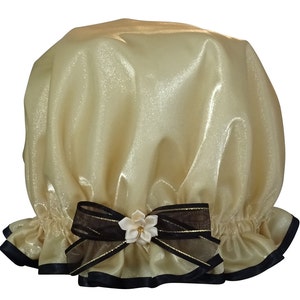 Può includere: Un cappello per la doccia in raso dorato con un nastro nero e oro e un accento di fiore bianco.