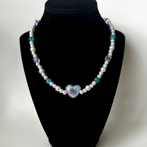 May include: A beaded necklace with a heart pendant. The necklace features white faux pearls, interspersed with teal and purple beads. The heart pendant is iridescent, reflecting multiple colors. The necklace is displayed on a black mannequin.