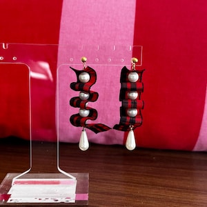 May include: A pair of statement earrings featuring red and black plaid ribbon, adorned with white faux pearls and a pear-shaped drop. The earrings are displayed on a clear acrylic stand against a pink and red striped background.