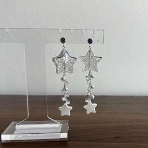 May include: A pair of silver star-shaped earrings with dangling white beads and a smaller white star at the bottom. The earrings are displayed on a clear acrylic stand against a neutral background.