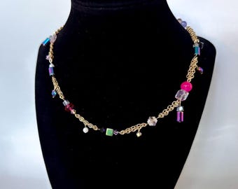 Op de afbeelding: Een gouden ketting met een verscheidenheid aan gekleurde kralen, waaronder roze, paars, groen en helder. De kralen zijn in verschillende vormen en maten, en sommige hebben een metallic afwerking.
