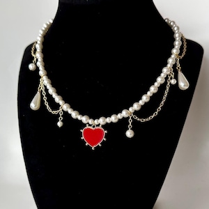May include: A white pearl necklace with a red heart pendant and gold chain accents.
