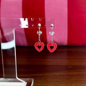 May include: A pair of red heart-shaped dangle earrings with clear crystal accents. The earrings are on a white display stand.