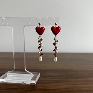 May include: A pair of dangling earrings with red heart-shaped pendants. Each earring features a cluster of small red and white beads, and a single white teardrop-shaped pearl. The earrings are displayed on a clear acrylic stand.