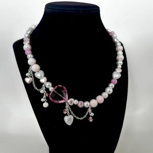 Puede incluir: Collar de cuentas con un colgante de corazón y abalorios. El collar presenta una mezcla de cuentas blancas, rosas y transparentes, con abalorios de corazón plateados colgando de la cadena. Se muestra sobre un maniquí negro.