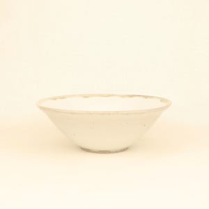 May include: A white ceramic bowl with a slightly flared shape. The bowl has a textured surface and a light beige rim. The bowl is empty and sits on a plain, off-white background. The bowl is likely for serving food.
