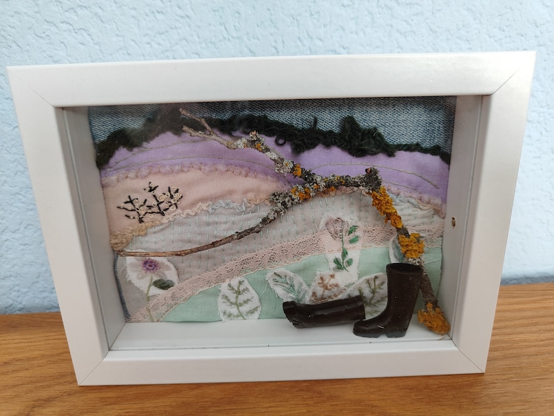 Peut inclure: Une &oelig;uvre d'art encadr&eacute;e repr&eacute;sentant une sc&egrave;ne de paysage avec une brindille, du lichen et des chutes de tissu dans des tons de rose, violet, vert et marron. Deux mini bottes en caoutchouc noires sont plac&eacute;es au bas du cadre.